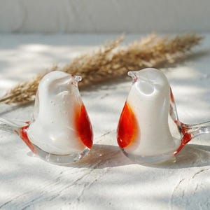May include: Two glass bird ornaments. Each bird is white with a red breast and tail feathers. The birds have clear glass wings and beaks. The ornaments are displayed on a white surface with a dried plant stem in the background.