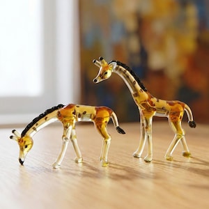 Könnte beinhalten: Zwei Giraffenfiguren aus Glas in bernsteinfarbenen und klaren Tönen mit dunkelbraunen Flecken und Mähnen. Eine Giraffe steht aufrecht mit erhobenem Kopf, während die andere in einer tieferen Haltung mit nach unten geneigtem Kopf positioniert ist.
