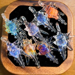 Könnte beinhalten: Eine Sammlung handgefertigter Glasanhänger in verschiedenen Farben, darunter Blau, Orange und schillernde Farbtöne. Jeder Anhänger hat ein einzigartiges, stacheliges Design und wird in einer Holzbox mit schwarzem Samtinterieur präsentiert.