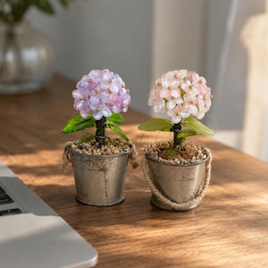 Può includere: Due piante decorative in vaso con grappoli di piccoli petali di pietra arrotondati in tonalità viola chiaro e rosa pallido. Ogni pianta ha foglie verdi, un gambo avvolto in filo metallico scuro ed è posta in un secchiello di metallo usurato con manico.