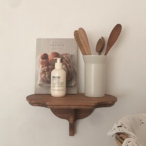 K&ouml;nnte beinhalten: Ein Holzregal mit geschwungener Front, das ein Buch, eine wei&szlig;e Lotionflasche und einen wei&szlig;en Keramikbeh&auml;lter mit Holzl&ouml;ffeln enth&auml;lt. Das Regal ist an einer wei&szlig;en Wand befestigt.