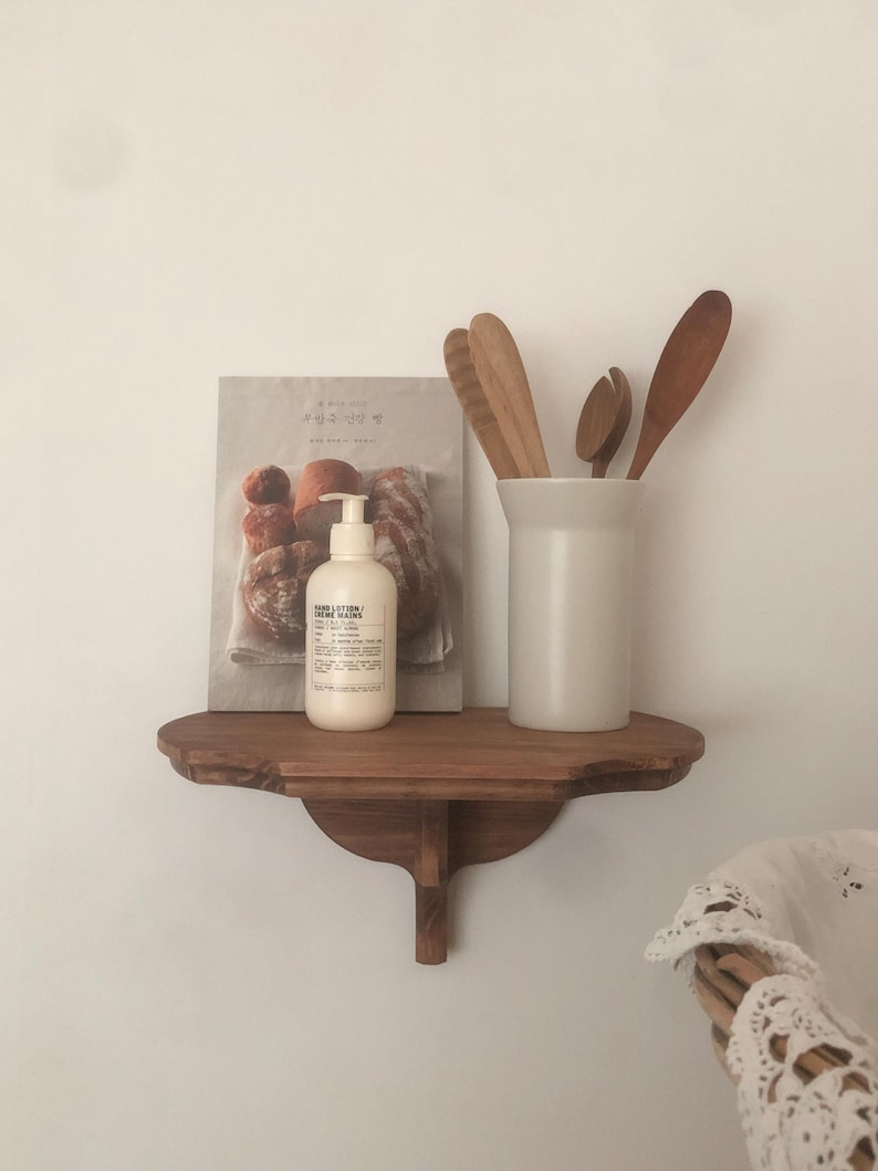 K&ouml;nnte beinhalten: Ein Holzregal mit geschwungener Front, das ein Buch, eine wei&szlig;e Lotionflasche und einen wei&szlig;en Keramikbeh&auml;lter mit Holzl&ouml;ffeln enth&auml;lt. Das Regal ist an einer wei&szlig;en Wand befestigt.
