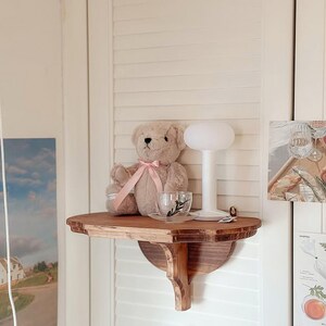 K&ouml;nnte beinhalten: Ein Holzregal mit einem Teddyb&auml;ren, einer Teetasse und einer kleinen Lampe. Der Teddyb&auml;r ist hellbraun mit einem rosa Band. Die Teetasse ist klar mit einem schwarzen Blattmuster. Die Lampe hat einen wei&szlig;en, abgerundeten Schirm.