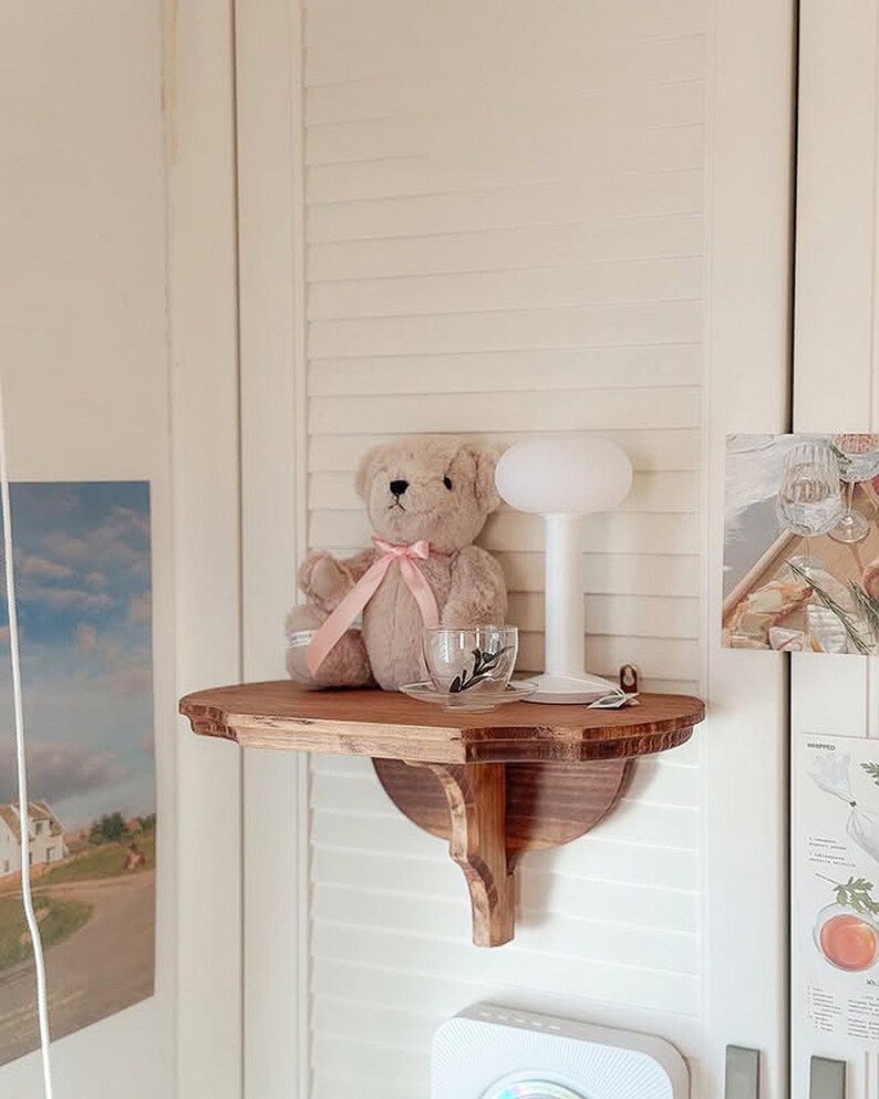 K&ouml;nnte beinhalten: Ein Holzregal mit einem Teddyb&auml;ren, einer Teetasse und einer kleinen Lampe. Der Teddyb&auml;r ist hellbraun mit einem rosa Band. Die Teetasse ist klar mit einem schwarzen Blattmuster. Die Lampe hat einen wei&szlig;en, abgerundeten Schirm.