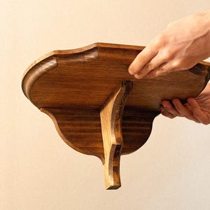 K&ouml;nnte beinhalten: Handgefertigtes Holzregal mit dekorativem, geschwungenem Design. Das Regal ist aus dunkel gebeiztem Holz gefertigt und hat eine gewellte Kante. Es wird von einer einzelnen, geschwungenen Halterung getragen. Zwei H&auml;nde halten das Regal.
