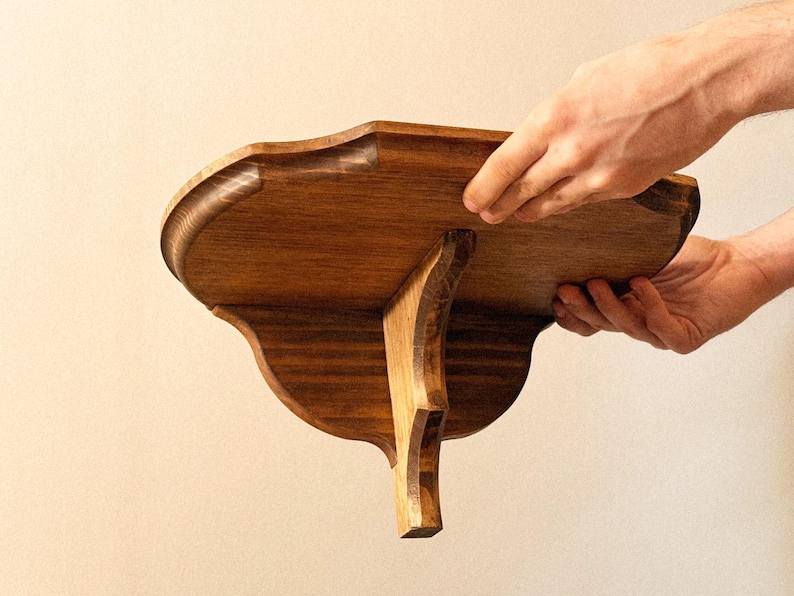 K&ouml;nnte beinhalten: Handgefertigtes Holzregal mit dekorativem, geschwungenem Design. Das Regal ist aus dunkel gebeiztem Holz gefertigt und hat eine gewellte Kante. Es wird von einer einzelnen, geschwungenen Halterung getragen. Zwei H&auml;nde halten das Regal.