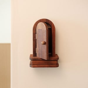May include: A miniature wooden door with an arched frame and a small knob. The door is slightly ajar, revealing a glimpse of the interior. The wood has a rich, dark brown color, and the piece is mounted on a small, rectangular base.