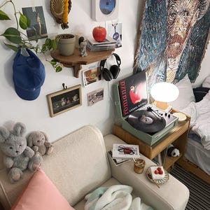 K&ouml;nnte beinhalten: Gem&uuml;tliches Zimmer mit wei&szlig;em Sofa, einem Nachttisch aus Holz mit Plattenspieler und einem Wandteppich von Van Gogh. Ein Regal zeigt eine Pflanze, einen roten Apfel und einen CD-Player. Pl&uuml;schhasen und ein rosa Kissen liegen auf dem Sofa.