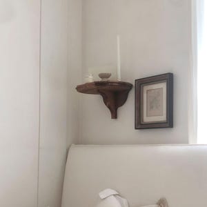 K&ouml;nnte beinhalten: Ein kleines, dunkles Holzregal an einer wei&szlig;en Wand, das eine kleine Schale, eine Kerze und ein Glas enth&auml;lt. Rechts h&auml;ngt ein gerahmtes Bild. Das Regal hat ein dekoratives, geschwungenes Design.
