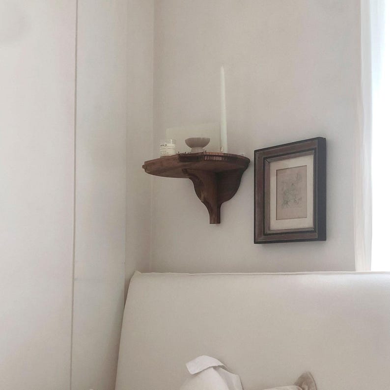 K&ouml;nnte beinhalten: Ein kleines, dunkles Holzregal an einer wei&szlig;en Wand, das eine kleine Schale, eine Kerze und ein Glas enth&auml;lt. Rechts h&auml;ngt ein gerahmtes Bild. Das Regal hat ein dekoratives, geschwungenes Design.