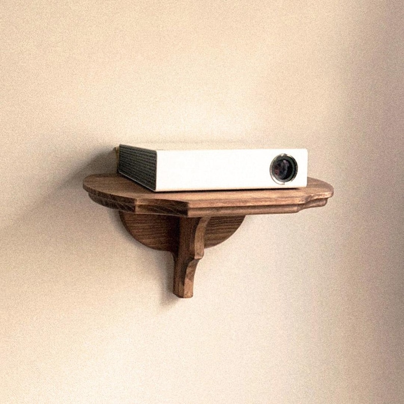 K&ouml;nnte beinhalten: Ein wei&szlig;er Projektor mit schwarzer Linse steht auf einem kleinen, runden Holzregal. Das Regal ist an einer beigefarbenen Wand befestigt. Das Regal hat ein dekoratives, geschwungenes Design und wird von einer einzelnen, verzierten Halterung getragen.