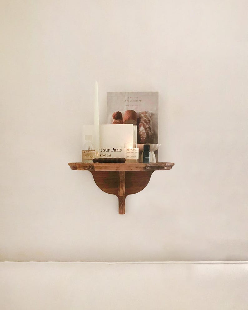 K&ouml;nnte beinhalten: Ein kleines, dunkles Holzregal an einer wei&szlig;en Wand, das eine Kerze, ein Buch und andere dekorative Gegenst&auml;nde enth&auml;lt. Das Buch hat Text auf dem Cover. Eine hohe, wei&szlig;e Kerze steht in einem Glasbeh&auml;lter. Das Regal hat ein geschwungenes Design.