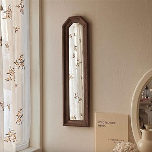 Può includere: Un lungo specchio rettangolare con cornice marrone scuro è appeso a una parete beige. Lo specchio riflette una tenda bianca trasparente con un motivo floreale. Un libro intitolato "Wild Flower Herbs" è visibile sotto.