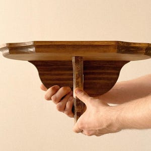 K&ouml;nnte beinhalten: Ein Regal aus dunkel gebeiztem Holz mit dekorativer Kante und Halterung. Das Regal wird gehalten. Es ist f&uuml;r die Wandmontage konzipiert. Die Abmessungen betragen ca. 30 cm Tiefe.