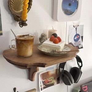 K&ouml;nnte beinhalten: Ein Holzregal mit einem Glas Eiskaffee, einem St&uuml;ck Kuchen mit Erdbeeren und einem schwarzen Kopfh&ouml;rer. Ein goldener Kerzenhalter und ein wei&szlig;er CD-Player sind an der Wand befestigt.