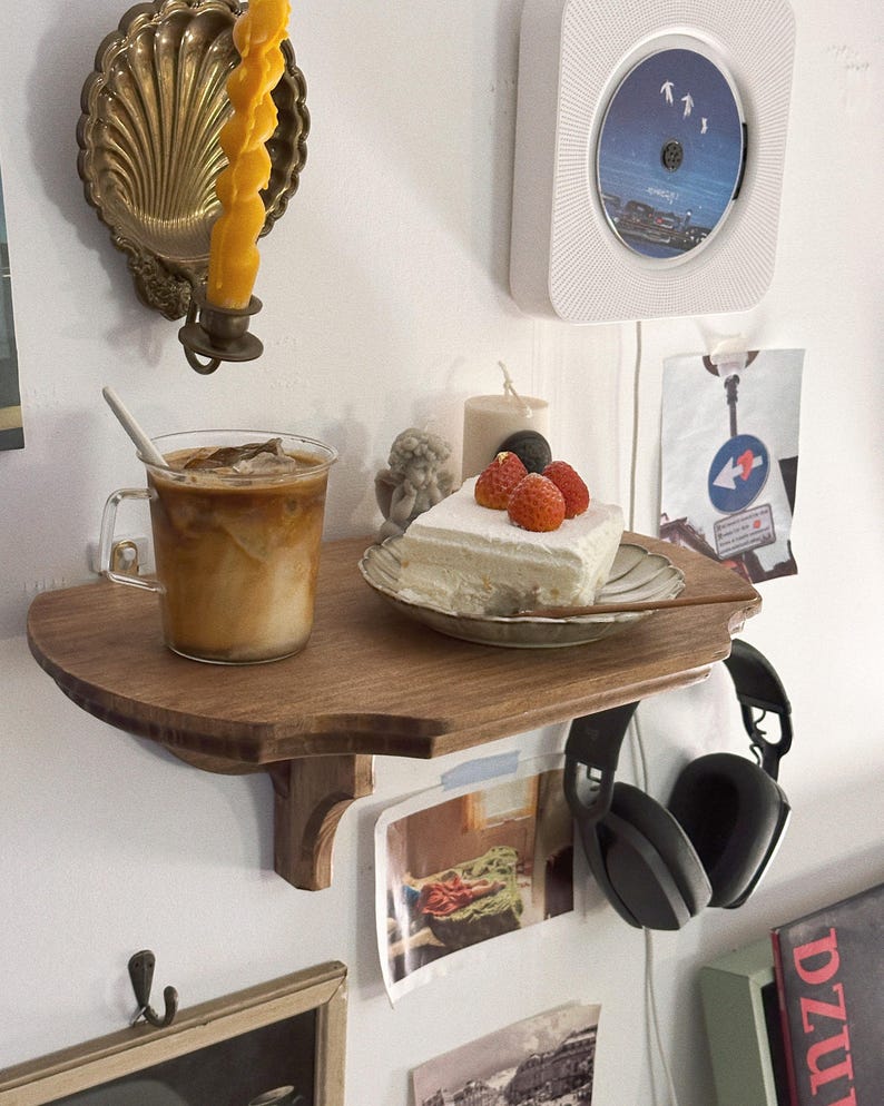 K&ouml;nnte beinhalten: Ein Holzregal mit einem Glas Eiskaffee, einem St&uuml;ck Kuchen mit Erdbeeren und einem schwarzen Kopfh&ouml;rer. Ein goldener Kerzenhalter und ein wei&szlig;er CD-Player sind an der Wand befestigt.