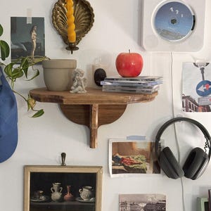 K&ouml;nnte beinhalten: Ein Holzregal zeigt einen roten Apfel, CDs, eine kleine Engelsfigur und eine Kerze. Ein gerahmtes Stillleben und ein an der Wand montierter CD-Player sind ebenfalls sichtbar. Ein goldfarbener Kerzenhalter mit einer gelben Kerze ist an der Wand befestigt.