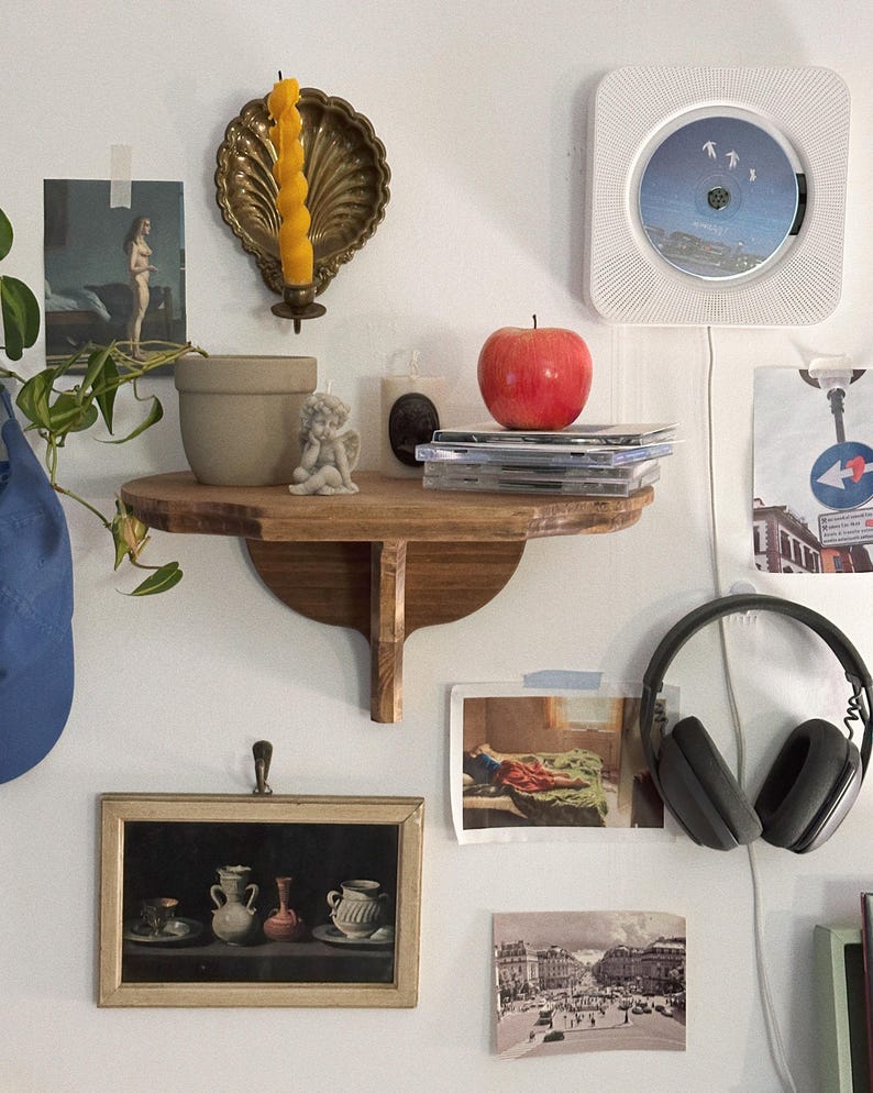 K&ouml;nnte beinhalten: Ein Holzregal zeigt einen roten Apfel, CDs, eine kleine Engelsfigur und eine Kerze. Ein gerahmtes Stillleben und ein an der Wand montierter CD-Player sind ebenfalls sichtbar. Ein goldfarbener Kerzenhalter mit einer gelben Kerze ist an der Wand befestigt.
