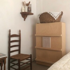 K&ouml;nnte beinhalten: Rustikales Schlafzimmer mit einem Holzstuhl mit Sprossenlehne und einem gewebten Sitz, einem hellen Holzschrank und einem Weidenkorb mit einem spitzenbesetzten Tuch. Ein kleines Regal zeigt dekorative Gegenst&auml;nde. Der Raum hat eine neutrale Farbpalette.