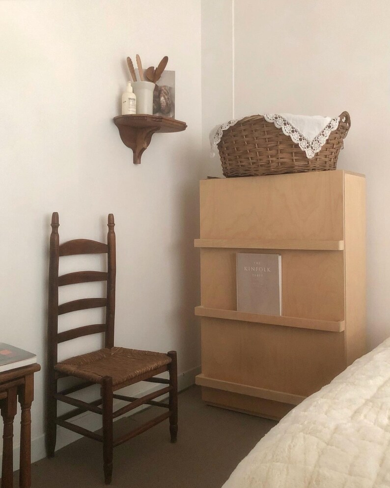 K&ouml;nnte beinhalten: Rustikales Schlafzimmer mit einem Holzstuhl mit Sprossenlehne und einem gewebten Sitz, einem hellen Holzschrank und einem Weidenkorb mit einem spitzenbesetzten Tuch. Ein kleines Regal zeigt dekorative Gegenst&auml;nde. Der Raum hat eine neutrale Farbpalette.