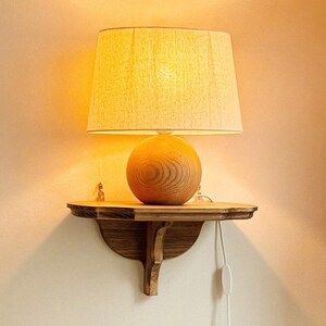 K&ouml;nnte beinhalten: Eine Lampe mit beigem Lampenschirm steht auf einem kleinen Regal aus dunklem Holz. Der Lampenfu&szlig; ist kugelf&ouml;rmig und aus Holz. Das Regal ist an einer hellen Wand befestigt. Ein wei&szlig;es Netzkabel mit Schalter ist sichtbar.