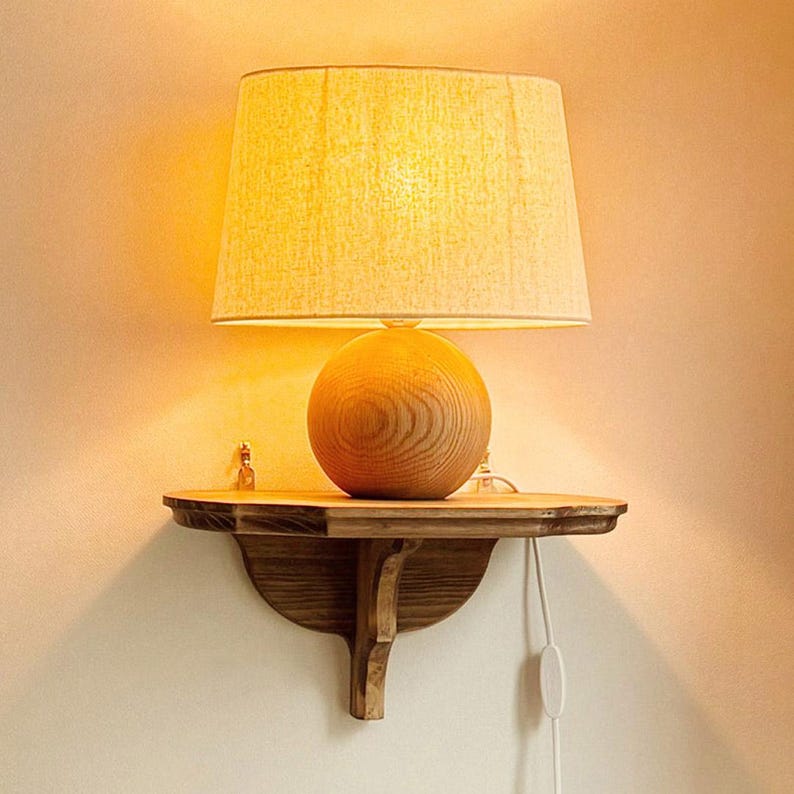 K&ouml;nnte beinhalten: Eine Lampe mit beigem Lampenschirm steht auf einem kleinen Regal aus dunklem Holz. Der Lampenfu&szlig; ist kugelf&ouml;rmig und aus Holz. Das Regal ist an einer hellen Wand befestigt. Ein wei&szlig;es Netzkabel mit Schalter ist sichtbar.