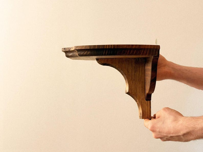K&ouml;nnte beinhalten: Ein handgefertigtes Holzregal mit dekorativem, geschwungenem Design. Das Regal wird von H&auml;nden gehalten, die seine reiche, nat&uuml;rliche Holzmaserung und warmen Braunt&ouml;ne zeigen. Perfekt zum Pr&auml;sentieren von Gegenst&auml;nden.