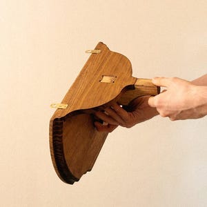 K&ouml;nnte beinhalten: Ein h&ouml;lzernes Wandregal mit geschwungenem Unterrand und Messingbeschl&auml;gen. Das Regal wird von den H&auml;nden einer Person vor einem neutralen Hintergrund gehalten. Das Regal ist aus hellbraunem Holz gefertigt.