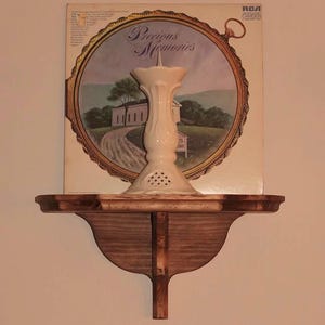 K&ouml;nnte beinhalten: Ein Holzregal mit dekorativer Halterung tr&auml;gt einen wei&szlig;en Kerzenhalter aus Keramik. Dahinter befindet sich ein Vintage-RCA-Schallplattenalbum mit dem Titel "Precious Memories" und einer Landschaftsszene. Das Regal ist an einer hellen Wand befestigt.