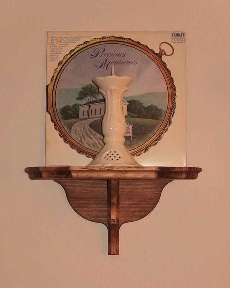 K&ouml;nnte beinhalten: Ein Holzregal mit dekorativer Halterung tr&auml;gt einen wei&szlig;en Kerzenhalter aus Keramik. Dahinter befindet sich ein Vintage-RCA-Schallplattenalbum mit dem Titel "Precious Memories" und einer Landschaftsszene. Das Regal ist an einer hellen Wand befestigt.