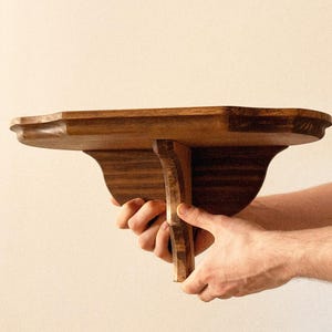 K&ouml;nnte beinhalten: Handgefertigtes Holzregal mit geschwungener Vorderkante und dekorativen St&uuml;tzen. Das Holz hat einen warmen, braunen Farbton. Das Regal ist f&uuml;r die Wandmontage konzipiert. Abmessungen ca. 30 cm tief.