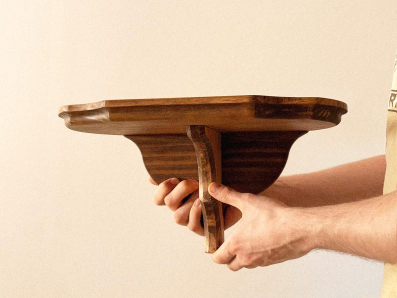 K&ouml;nnte beinhalten: Handgefertigtes Holzregal mit geschwungener Vorderkante und dekorativen St&uuml;tzen. Das Holz hat einen warmen, braunen Farbton. Das Regal ist f&uuml;r die Wandmontage konzipiert. Abmessungen ca. 30 cm tief.