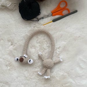 May include: A handmade beige crochet giraffe toy with white accents. The giraffe has a long neck, small arms and legs, and large white-rimmed eyes. Crochet supplies, including yarn, scissors, and a crochet hook, are visible in the background.