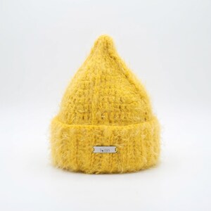 May include: A bright yellow beanie with a fuzzy, textured knit construction and a pointed crown. The hat features a folded brim and a small, rectangular silver-toned metal tag centered on the front with the text T BB engraved on its surface.