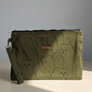 Könnte beinhalten: Olivgrüne Clutch mit einem schwarzen Katzenmuster. Die rechteckige Tasche hat einen schwarzen Reißverschluss und eine passende Handschlaufe. Ein kleines, rechteckiges Lederetikett befindet sich mittig auf der Vorderseite.