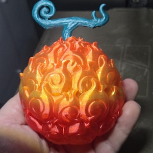 May include: A 3D-printed fruit shaped like flames, with a blue stem. The fruit's colours shift from red at the bottom to orange and yellow at the top, mimicking fire. The stem has a swirled design. The object is held in a hand.