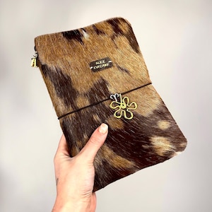 Può includere: Pochette rettangolare con motivo animalier marrone e beige. La pochette ha un elastico nero con un ciondolo a forma di fiore dorato. Il testo "ALICE EVELASE" è su una piccola etichetta rettangolare. È visibile un tirazip dorato.