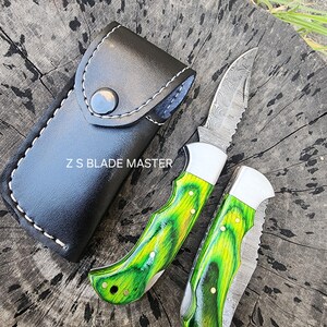 May include: Two folding knives with green and yellow handles and patterned blades, next to a black leather sheath. The blades have a serrated edge. The text "Z S BLADE MASTER" is visible on the sheath.