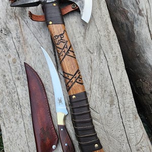 May include: Axe and knife set with a skull and crossbones design. The axe has a wooden handle with carved details and a leather grip. The knife has a wooden handle and a leather sheath. The set is displayed on a wooden surface.