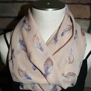 Puede incluir: Bufanda infinita de color melocotón con un estampado de plumas repetido en tonos grises, morados y rosas. La bufanda está drapeada alrededor del cuello de un maniquí, mostrando su diseño y textura. La bufanda está hecha de un material ligero.