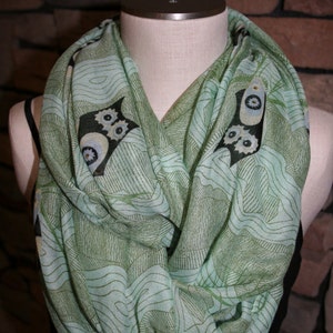 May include: A green and black patterned infinity scarf with a design of stylized owls. The scarf is made of a soft, lightweight fabric and is perfect for adding a touch of style to any outfit.