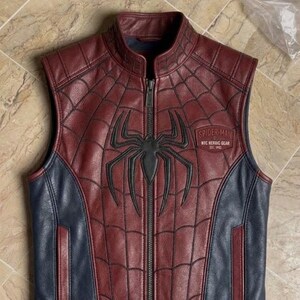 May include: A sleeveless leather vest in maroon and navy blue featuring a black spider emblem on the chest. The maroon panels have a black web pattern. A patch on the chest reads Spider-Man NYC Nerdic Gear Est 1962. It has a front zipper closure.