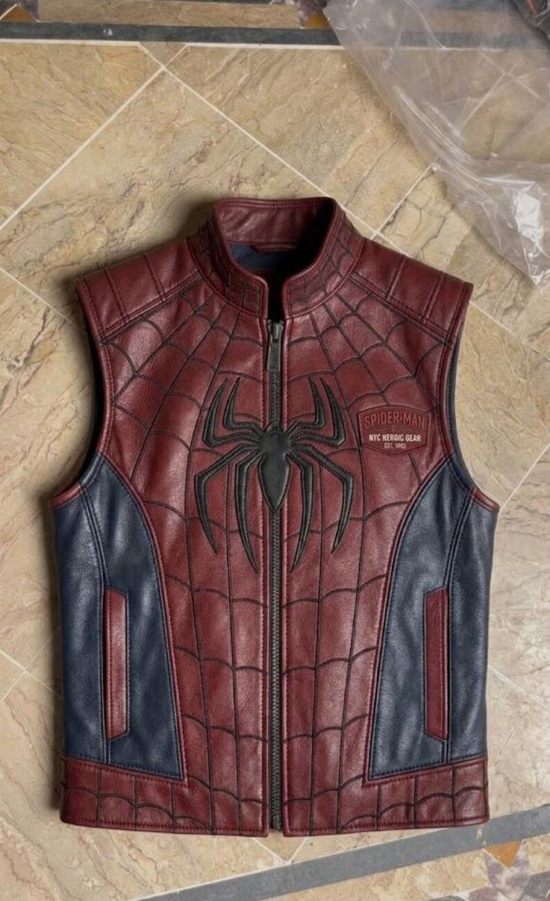 May include: A sleeveless leather vest in maroon and navy blue featuring a black spider emblem on the chest. The maroon panels have a black web pattern. A patch on the chest reads Spider-Man NYC Nerdic Gear Est 1962. It has a front zipper closure.