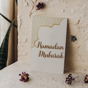 May include: A white card with gold filigree and the words "Ramadan Mubarak" in elegant script. The card is set against a textured, off-white backdrop, adorned with dried flowers, creating a festive and elegant aesthetic.