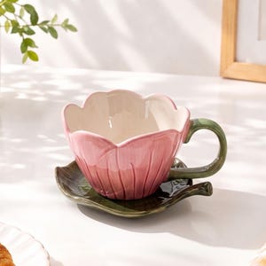 Puede incluir: Una taza de té de cerámica con forma de tulipán rosa y un asa en forma de tallo verde, apoyada sobre un plato a juego diseñado como una hoja de color verde oscuro. La taza tiene un acabado liso y brillante con un interior color crema.
