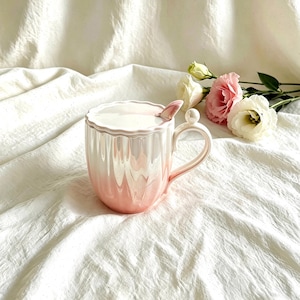 May include: A pink and white ceramic mug with a matching lid and spoon. The mug has a pearlescent finish and a scalloped rim. The handle features a small decorative ball. The mug is on a white, textured surface.