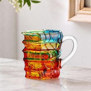 Op de afbeelding: Een vierkante glazen mok met een ontwerp van gestapelde boeken in levendige, doorschijnende kleuren zoals blauw, groen, geel, oranje en rood. De mok heeft een helder, gebogen handvat en een getextureerd oppervlak dat boekruggen nabootst.