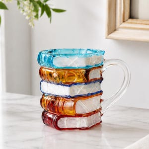 May include: A glass mug shaped like a stack of five books with a clear handle. The books are colored in shades of blue, amber, and red, featuring textured white page edges. The mug has a cylindrical body and a smooth, transparent glass handle.
