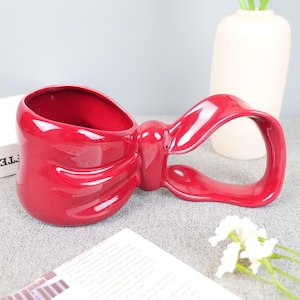 May include: A glossy, red ceramic mug shaped like a bow. The mug has a large opening and a handle formed by the bow's loop. The design is smooth and modern, suitable for beverages or as a decorative piece.