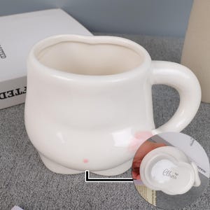 May include: A white ceramic mug with a rounded body and a large handle. The mug has a unique shape with a small base and a wide opening. The name "Ethan" is printed on a small, white lid. The mug is likely used for drinking beverages.